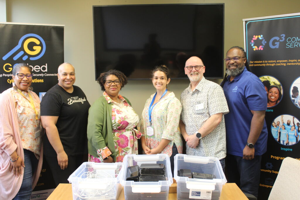 Six adults standing in a conference room in front of a table with plastic bins on top containing cell phones.
