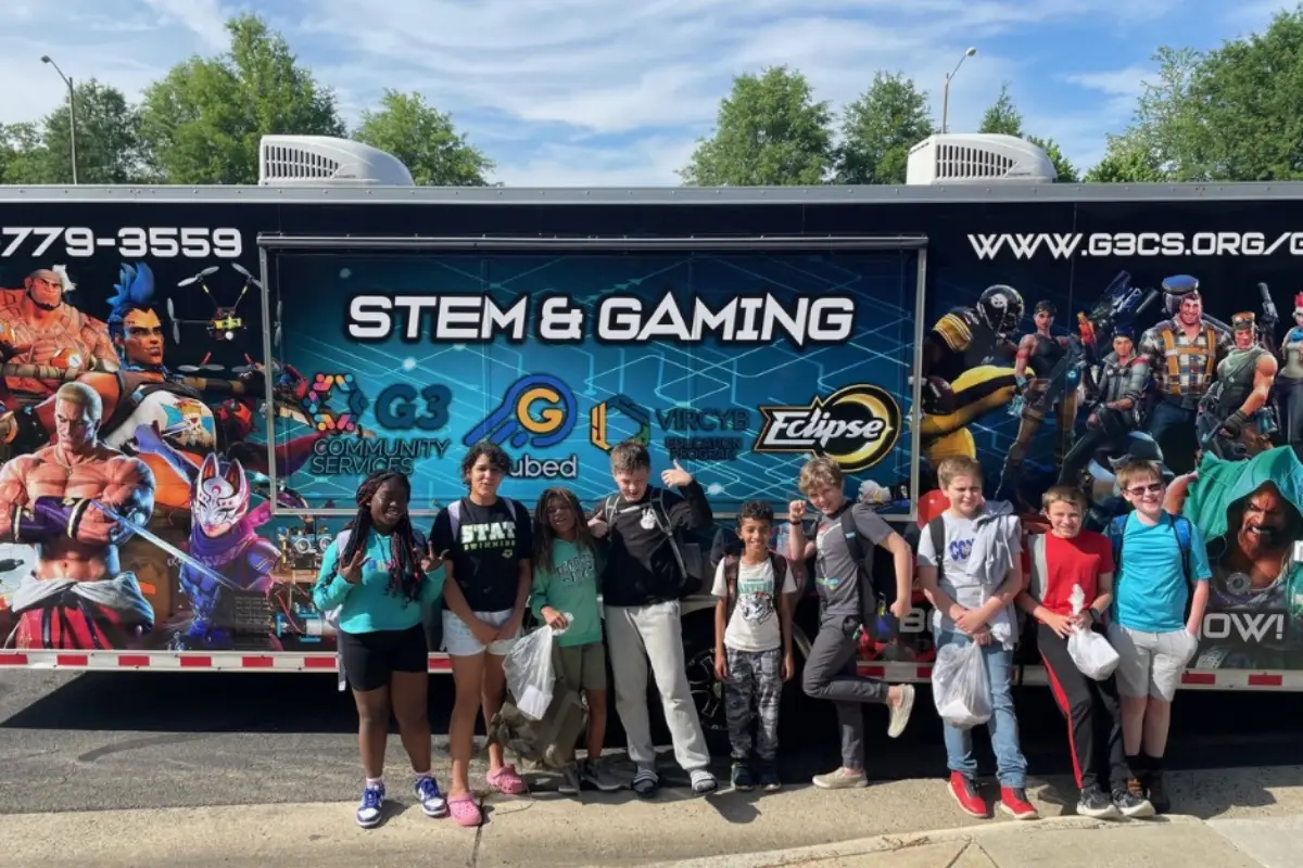 A group of children standing in front of a mobile video game truck with characters pictured on the outside and the words "STEM and Gaming" in the middle.