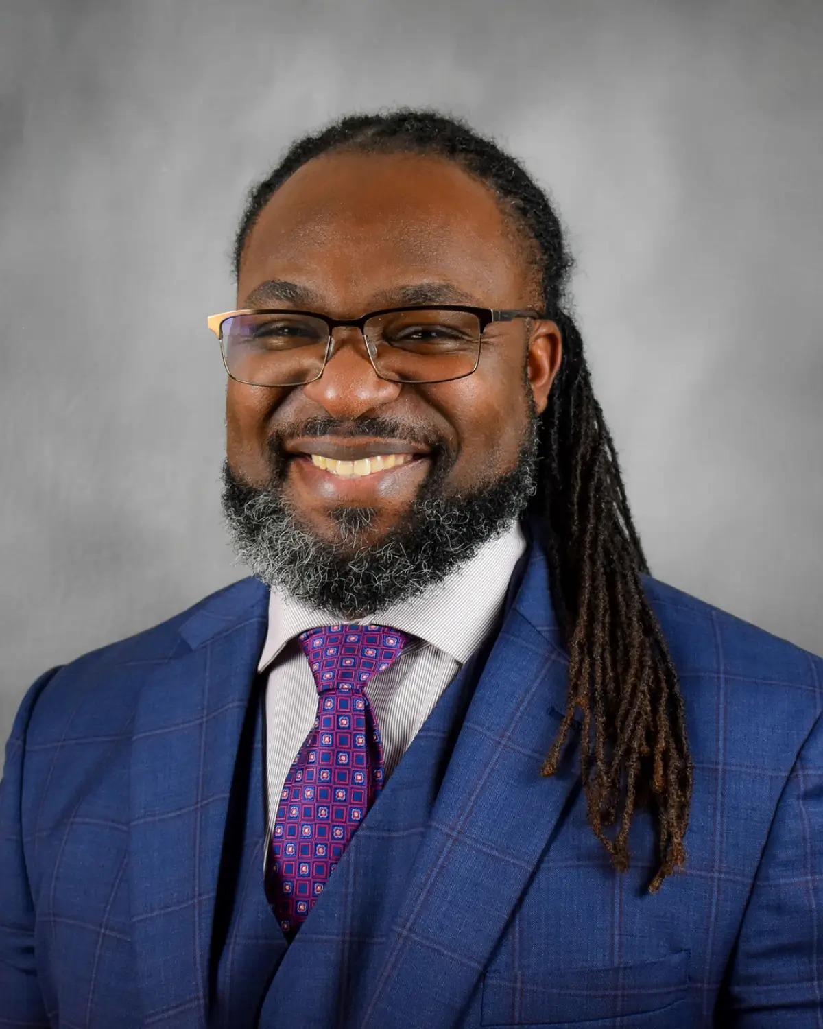 A man in a blue suit with a white shirt and purple tie smiling in front of a light gray background.